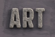 The word 'ART' is carved in a textured, concrete-like material, with rough, brush-stroke patterns covering the surface. It is set against a slate grey, wooden textured background.
