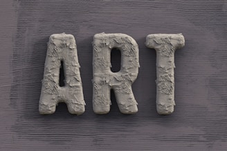 The word 'ART' is carved in a textured, concrete-like material, with rough, brush-stroke patterns covering the surface. It is set against a slate grey, wooden textured background.