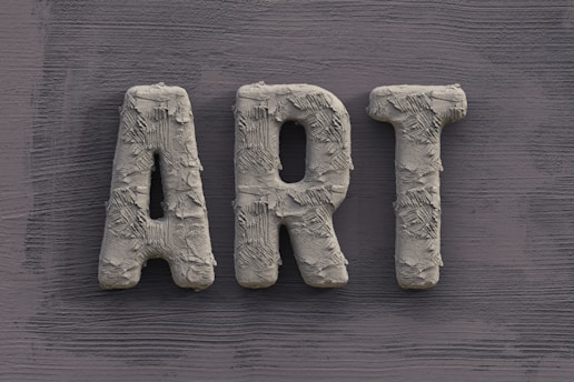 The word 'ART' is carved in a textured, concrete-like material, with rough, brush-stroke patterns covering the surface. It is set against a slate grey, wooden textured background.