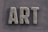 The word 'ART' is carved in a textured, concrete-like material, with rough, brush-stroke patterns covering the surface. It is set against a slate grey, wooden textured background.