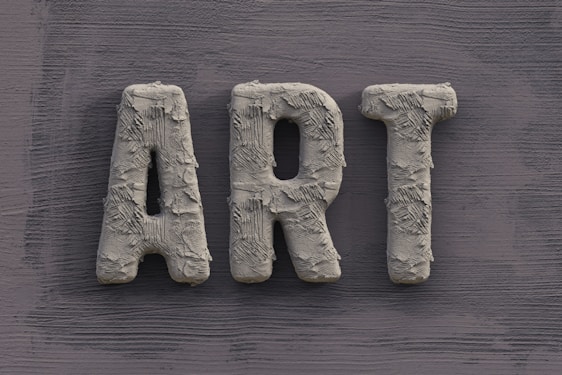 The word 'ART' is carved in a textured, concrete-like material, with rough, brush-stroke patterns covering the surface. It is set against a slate grey, wooden textured background.