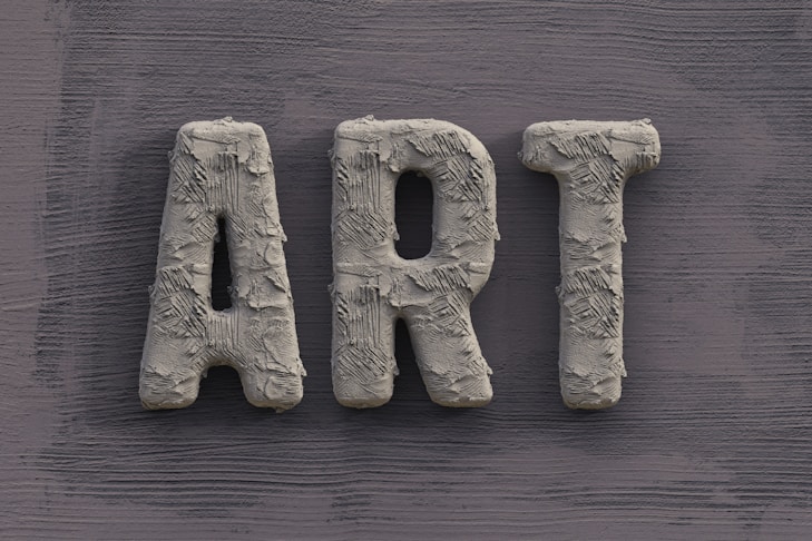 The word 'ART' is carved in a textured, concrete-like material, with rough, brush-stroke patterns covering the surface. It is set against a slate grey, wooden textured background.