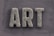 The word 'ART' is carved in a textured, concrete-like material, with rough, brush-stroke patterns covering the surface. It is set against a slate grey, wooden textured background.
