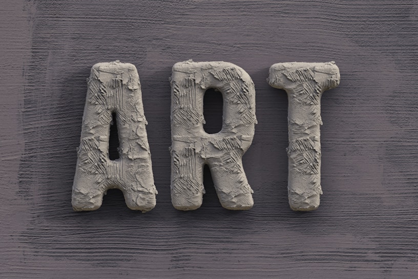 The word 'ART' is carved in a textured, concrete-like material, with rough, brush-stroke patterns covering the surface. It is set against a slate grey, wooden textured background.