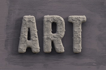 The word 'ART' is carved in a textured, concrete-like material, with rough, brush-stroke patterns covering the surface. It is set against a slate grey, wooden textured background.
