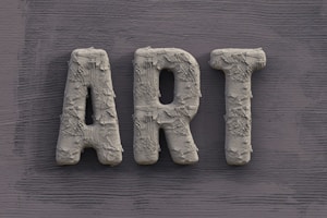 The word 'ART' is carved in a textured, concrete-like material, with rough, brush-stroke patterns covering the surface. It is set against a slate grey, wooden textured background.
