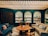 A cozy room features a round wooden coffee table surrounded by an assortment of chairs and couches with various cushions. The back wall showcases three arched shelves filled with vinyl records or book covers. The ceiling is designed with wooden slats, and the floor has a striped black and white pattern. The room is warmly lit, enhancing the welcoming atmosphere.