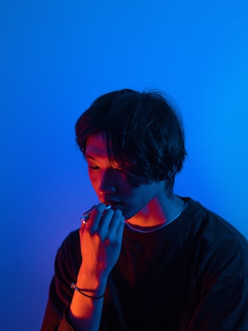 A person is seen in a dimly lit environment with contrasting blue and red lighting creating a dramatic effect. The individual appears contemplative or introspective, with their hand raised near their mouth.
