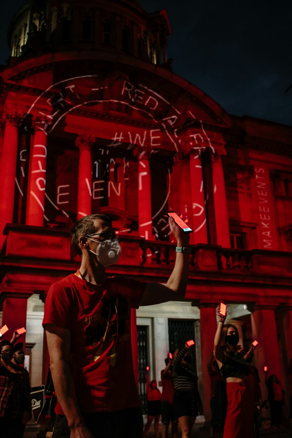 검은 고글을 쓴 흰색 크루넥 티셔츠를 입은 남자가 밤에 빨간 건물 근처에 서 있다