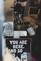 A person operates a coffee machine, with two white mugs placed under it. Below the machine, a sign reads 'You are here, and so is ...' with an advertisement for Oatly products. The setting has a rustic or cozy vibe with visible metallic textures.