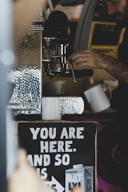 A person operates a coffee machine, with two white mugs placed under it. Below the machine, a sign reads 'You are here, and so is ...' with an advertisement for Oatly products. The setting has a rustic or cozy vibe with visible metallic textures.