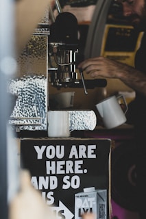 A person operates a coffee machine, with two white mugs placed under it. Below the machine, a sign reads 'You are here, and so is ...' with an advertisement for Oatly products. The setting has a rustic or cozy vibe with visible metallic textures.