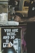 The image depicts a person operating a coffee machine with a focus on their hands holding a cup. There is a prominent poster in the foreground with the message 'YOU ARE HERE. AND SO IS THIS,' accompanied by an image of a carton labeled 'OATLY!' The background contains blurry elements of a coffee shop setting.