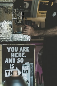 The image depicts a person operating a coffee machine with a focus on their hands holding a cup. There is a prominent poster in the foreground with the message 'YOU ARE HERE. AND SO IS THIS,' accompanied by an image of a carton labeled 'OATLY!' The background contains blurry elements of a coffee shop setting.
