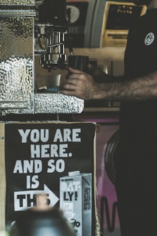The image depicts a person operating a coffee machine with a focus on their hands holding a cup. There is a prominent poster in the foreground with the message 'YOU ARE HERE. AND SO IS THIS,' accompanied by an image of a carton labeled 'OATLY!' The background contains blurry elements of a coffee shop setting.