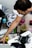 A man reaches into a plastic container with various art supplies scattered around, including spray paint cans, paint buckets, brushes, and a respirator mask. His arm and hand are visible as he interacts with the materials, which are placed on a rough surface.