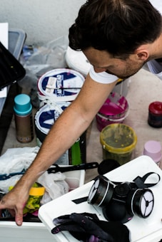 A man reaches into a plastic container with various art supplies scattered around, including spray paint cans, paint buckets, brushes, and a respirator mask. His arm and hand are visible as he interacts with the materials, which are placed on a rough surface.