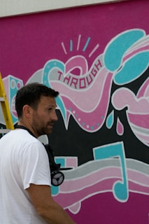 A man stands beside a vibrant mural featuring abstract shapes and musical notes in pink, teal, and black. The word 'THROUGH' is incorporated into the design. The mural includes various patterns and elements such as polka dots and dripping shapes. A ladder is partially visible, and the man appears to be holding spray paint gear.