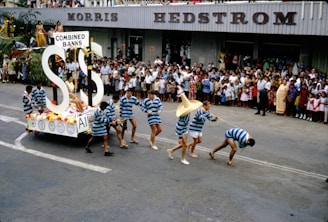 A parade scene featuring a float with large dollar signs and the words 'Combined Banks.' Participants in striped costumes are seen pulling the float, while a large crowd of spectators, including families and children, watch from the sidelines. The event takes place on a street in front of a building with the signage 'Morris Hedstrom.'
