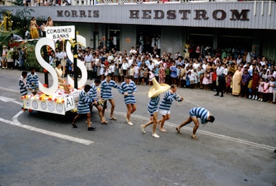 A parade scene featuring a float with large dollar signs and the words 'Combined Banks.' Participants in striped costumes are seen pulling the float, while a large crowd of spectators, including families and children, watch from the sidelines. The event takes place on a street in front of a building with the signage 'Morris Hedstrom.'