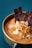 A close-up photograph of a bowl containing creamy soup or puree, garnished with thin, crispy slices of garlic and strips of dark, dried meat. The dish is set against a teal fabric background, creating a contrast with the warm tones of the food.