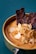 A close-up photograph of a bowl containing creamy soup or puree, garnished with thin, crispy slices of garlic and strips of dark, dried meat. The dish is set against a teal fabric background, creating a contrast with the warm tones of the food.