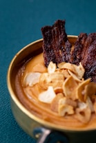 A close-up photograph of a bowl containing creamy soup or puree, garnished with thin, crispy slices of garlic and strips of dark, dried meat. The dish is set against a teal fabric background, creating a contrast with the warm tones of the food.