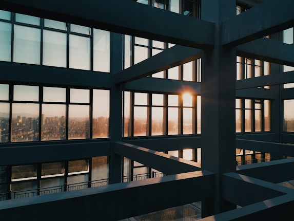 A modern architectural structure with large grid-like windows, capturing the sunset. The sunlight casts a warm glow through the windows, illuminating the stark lines and angles of the building's interior framing the distant cityscape outside.