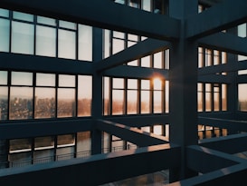 A modern architectural structure with large grid-like windows, capturing the sunset. The sunlight casts a warm glow through the windows, illuminating the stark lines and angles of the building's interior framing the distant cityscape outside.
