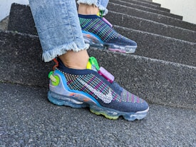 A pair of colorful, modern sneakers with multi-colored detailing and transparent soles is worn on a person's feet. The sneakers feature a prominent brand logo and are paired with light blue jeans with frayed hems. The person is standing on a set of rough-textured, concrete steps, suggesting an urban environment.