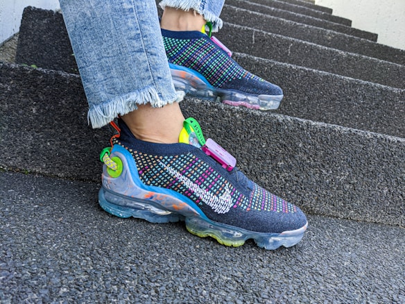 A pair of colorful, modern sneakers with multi-colored detailing and transparent soles is worn on a person's feet. The sneakers feature a prominent brand logo and are paired with light blue jeans with frayed hems. The person is standing on a set of rough-textured, concrete steps, suggesting an urban environment.