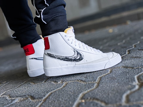 A close-up shot of a person's feet wearing white high-top sneakers with a black swoosh design. The sneakers have a textured leather finish and red accent on the back. The person is standing on a cobblestone pavement, wearing dark pants.