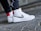 A close-up shot of a person's feet wearing white high-top sneakers with a black swoosh design. The sneakers have a textured leather finish and red accent on the back. The person is standing on a cobblestone pavement, wearing dark pants.