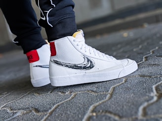 A close-up shot of a person's feet wearing white high-top sneakers with a black swoosh design. The sneakers have a textured leather finish and red accent on the back. The person is standing on a cobblestone pavement, wearing dark pants.