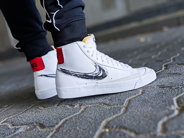 A close-up shot of a person's feet wearing white high-top sneakers with a black swoosh design. The sneakers have a textured leather finish and red accent on the back. The person is standing on a cobblestone pavement, wearing dark pants.