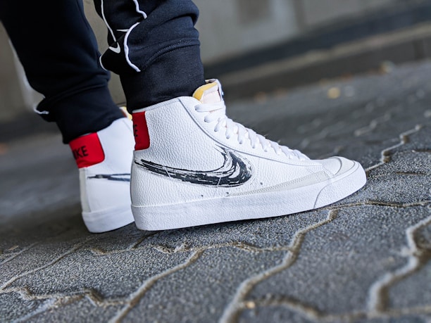 A close-up shot of a person's feet wearing white high-top sneakers with a black swoosh design. The sneakers have a textured leather finish and red accent on the back. The person is standing on a cobblestone pavement, wearing dark pants.