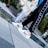 A person wearing stylish white sneakers with a metallic swoosh design stands by a fountain. The sneakers are accented with red and yellow details, and the person is dressed in black pants with a repeated white Nike swoosh pattern on the side. The background features an outdoor urban setting with stone tiles and greenery.