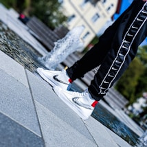 A person wearing stylish white sneakers with a metallic swoosh design stands by a fountain. The sneakers are accented with red and yellow details, and the person is dressed in black pants with a repeated white Nike swoosh pattern on the side. The background features an outdoor urban setting with stone tiles and greenery.