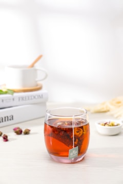 A transparent glass filled with a rich, amber-colored tea, featuring a visible tea bag with dried floral elements. Accompanying the glass are scattered dried rose buds and a small bowl with mixed dried flowers. In the background, a white ceramic mug with a wooden spoon rests on top of two stacked books titled 'FREEDOM'. The setting is minimalistic and serene.