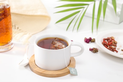 A white cup filled with tea sits on a wooden coaster, accompanied by a teabag whose tag is visible over the side. Nearby, a saucer holds a variety of dried herbs and berries, and a glass of amber-colored iced tea rests on the surface. A light fabric and a green leaf add to the serene and refreshing setting.