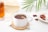 A white cup filled with tea sits on a wooden coaster, accompanied by a teabag whose tag is visible over the side. Nearby, a saucer holds a variety of dried herbs and berries, and a glass of amber-colored iced tea rests on the surface. A light fabric and a green leaf add to the serene and refreshing setting.