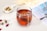 A clear glass cup filled with reddish herbal tea, featuring a tea bag inside. Around the glass cup, there are dry flower petals and buds scattered on the light-colored surface. To the left, there's a small white bowl containing a variety of dried herbs and flowers. A blurred, partially visible magazine or book with a photo of a palm tree provides a soft background, along with some light-colored fabric.