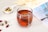 A clear glass cup filled with reddish herbal tea, featuring a tea bag inside. Around the glass cup, there are dry flower petals and buds scattered on the light-colored surface. To the left, there's a small white bowl containing a variety of dried herbs and flowers. A blurred, partially visible magazine or book with a photo of a palm tree provides a soft background, along with some light-colored fabric.