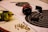 Target sheets with concentric circles are laid out on a table, accompanied by two sets of ear protection earmuffs and a small pile of bullet casings. One set of earmuffs is black and red, while the other is yellow and black. A black object, possibly a magazine or stand, is placed on one of the target sheets.