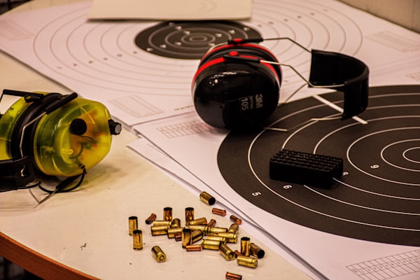 Target sheets with concentric circles are laid out on a table, accompanied by two sets of ear protection earmuffs and a small pile of bullet casings. One set of earmuffs is black and red, while the other is yellow and black. A black object, possibly a magazine or stand, is placed on one of the target sheets.
