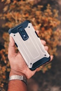 A hand holding a smartphone with a rugged, metallic case. The smartphone has a dual camera setup and is held against a blurred background of autumnal foliage.