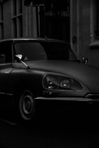 A vintage car is parked in a dimly lit urban setting, primarily captured in shadows and low light, emphasizing the vehicle's sleek design and classic features.