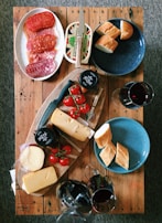A wooden table is laid with an assortment of food items including sliced cured meats, blocks of cheese, cherry tomatoes on the vine, and bread rolls. There are two glasses and a decanter of red wine. A knife rests on a block of cheese, and there are colorful plates placed on the table.