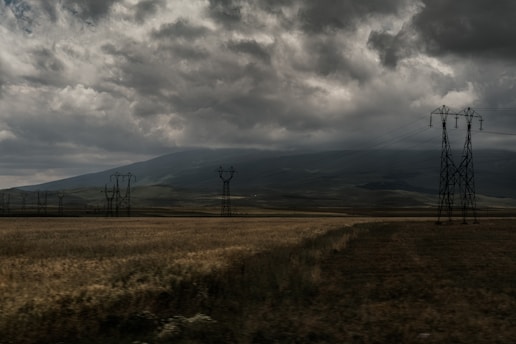 A vast field stretches into the distance with tall electrical transmission towers standing throughout. Dark, ominous clouds cover the sky, casting shadows over the landscape. Distant hills are partially visible through the mist.