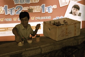 A person is arranging small potted plants on a ledge. Beside them, there is a larger box containing more plants. The background features a brightly colored advertisement with large text in a foreign language and an image of a surprised child holding a phone.